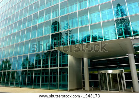 The deserted front door of a large commercial office block, surrounded by trees reflected in the glass. One tree reflection appears to grow out of a pillar of the entrance.