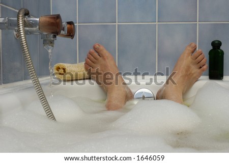 A man\'s feet, poking out of a bubble bath, with water running in to the side. A loofah and bottle of bath lotion to either side. Space for text on the blue tiled wall above the feet.