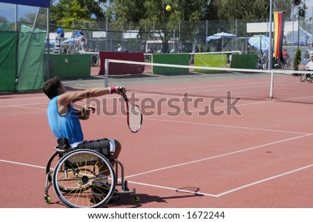 A wheelchair tennis player during a tennis championship match, serving. The ball, with motion blur, is visible centre top.