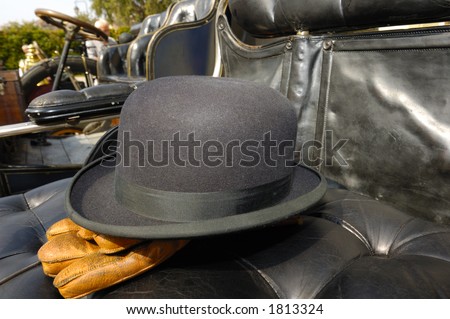 The black leather seat of a vintage motor-car with the driver\'s bowler hat and brown leather gloves placed carefully on it. Other vintage cars, lined up, can be seen in the background.