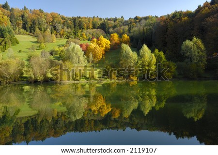 The colours of autumn in  the early-morning light, with the trees reflected in the waters of a lake. A tiny figure can be seen walking past the brightest trees.