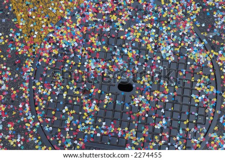 Confetti scattered across the metal cover of a drain in the street, the random scattering of the confetti contrasting with the regular grid on the manhole cover.