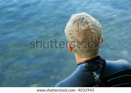 A triathlon swimmer, dressed in a wet suit, psyching himself up to go as he gazes at the cold clear water.