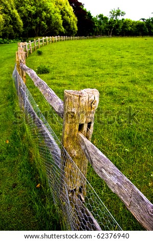 View of a green colourful field with wooden fence running through the middle with trees in the background