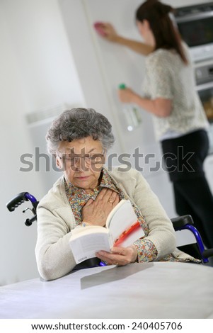 Elderly woman reading book, home helper in background