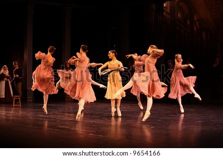 group of classic ballet female dancers dressed pink dancing around female dancer dressed white holding mandolin lit by stage lights