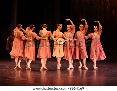 group of classic ballet female dancers dressed pink dancing at row while center dancer dressed white holding mandolin lit by stage lights