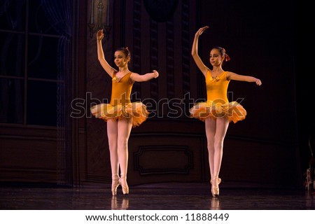two classic ballet girl dancers in yellow dresses on stage standing with raised hands lit by stage light