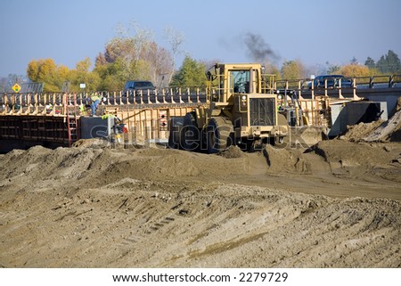 Workmen adjust concrete forms for bridge while heavy equipment pushes dirt