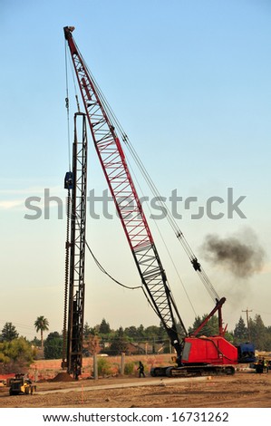 A power auger to drill deep holes is supported by a crane on a construction job site