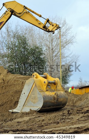Digging bucket rests detached from crane on mound of dirt while crane is used to lift heavy load