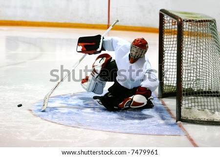 Goalie in generic white jersey with generic red goalie mask makes a tremendous diving attempt to prevent the puck (frozen in flight) from entering the goal