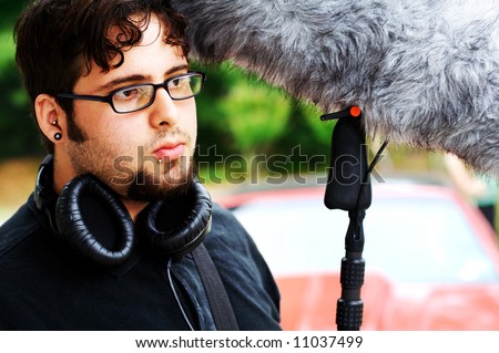 A sound man on a movie set holding a boom mic with a wind sock on it and wearing headphones.