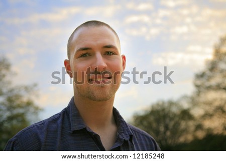 A young man outside under a beautiful sky. He is smiling and looking into the camera.