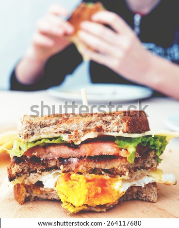 Bitten egg sandwich and person eating in the background. Image filtered