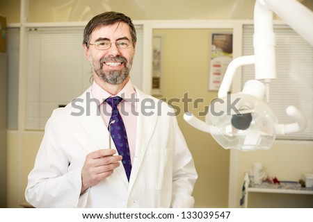 Portrait of happy middle-aged dentist in his cabinet, wearing lab coat, dentist mirror in his hand