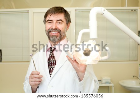 Portrait of happy middle-aged dentist in his cabinet, wearing lab coat, dentist mirror in his hand