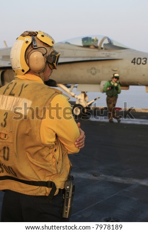 An Aircraft Director (Yellow Shirt) Awaits the Launch of an F/A-18C Hornet on a Nuclear Aircraft Carrier, as a Catapult Crewmember (Green Shirt) Looks on