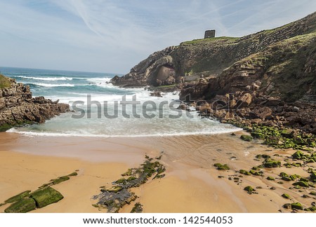 Santa Justa beach located on the coast of Cantabria to the north of Spain, they are some rocks in the sand, and the hills on either side, on a hillside is a hermitage