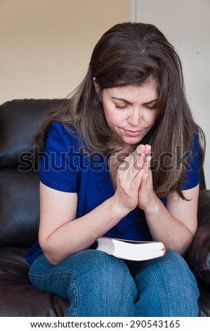 Christian Daily Devotional: Woman praying with the holy bible on his knees folded her hands in prayer while sitting on the couch in the room
