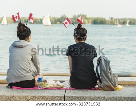 TORONTO,CANADA-JULY 1,2015: Canada\'s day celebrations, friends enjoying a day out in the Toronto Harborfront in Canada Day.Two girls sitting and eat on the beach with Canada flag on his head.