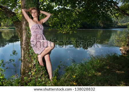 Beautiful young woman leaning on a tree by the river