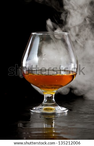 dramatic shot of a brandy snifter with whiskey served on a dark bar with fog around it