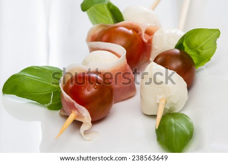 close up of a fresh mozzarella and tomato appetizer with basil and proscuitto on a white plate