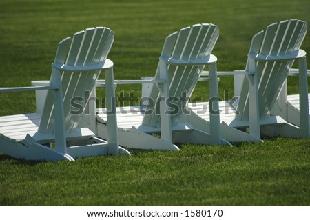 A row of three white, wooden  lawn chairs on a vividly green lawn.