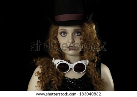 Beautiful young woman with long, curly, red hair wearing top hat, vintage sequin tank top, and goggles on black background.