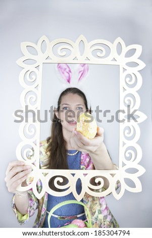 Pretty young woman wearing rabbit ears and a colorful jacket with long hair holding yellow sequin Easter egg through wooden picture frame.