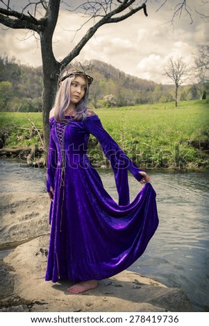 Woman dressed in purple velvet gown and twig crown standing on a rock by a creek,