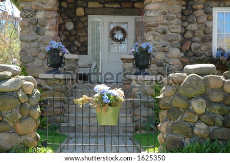 Home entrance to cobblestone house accented by purple blue hydrangea flowers in urns and a hanging pot on a gate