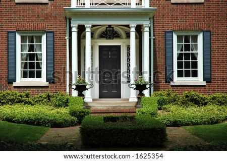 Red brick house with black shutters and green shrubs with front door flanked by pillars and two flower iron urns; luxury home / house