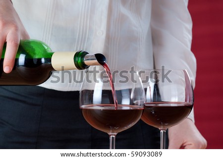 Women pouring red wine into wineglass. Next to her stay two wineglasses and carafe. Red background behind her with sun beams and shades.