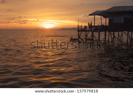 Sunset over calm ocean horizon with house on stilts with people in silhouette