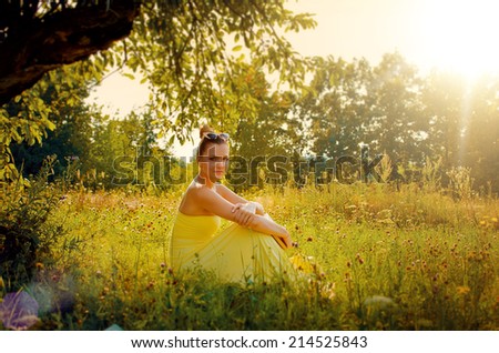 Beautiful slim woman in yellow dress sitting on the grass in the garden at sunset