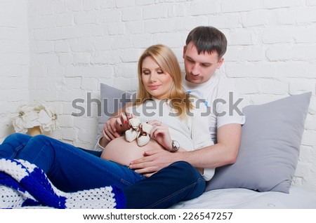 Pregnant woman and her husband holding booties. Prospective parents lie on the couch.
