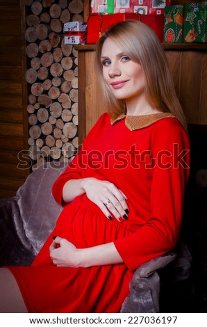 Happy pregnant woman in a red dress sitting by the fireplace with lots of gifts