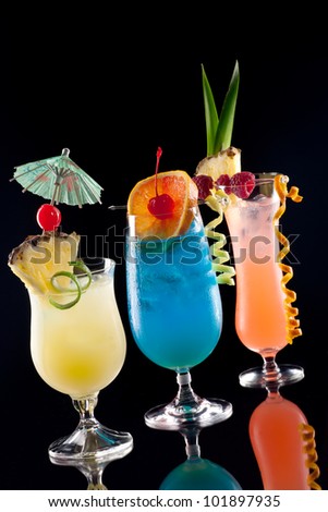 Rum Runner, Bahama Mama, and Blue Lagoon cocktaisl over black background on reflection surface, garnished with pineapple flag, fresh raspberry, maraschino cherry, and lime twist.