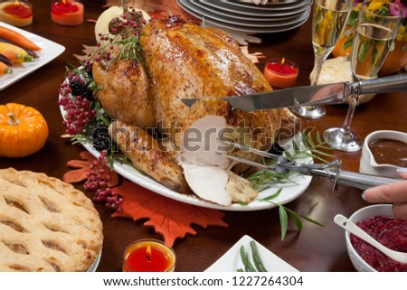 Carving roasted pepper turkey for Thanksgiving, garnished with pink pepper, blackberry, and fresh rosemary twigs on a dinner table decorated with mini pumpkins, beans, carrots, baked potato, and flute