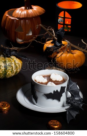 Cup of hot chocolate with ghost marshmallow, surrounded with pumpkins, candles, and Halloween decoration. Halloween drinks series.