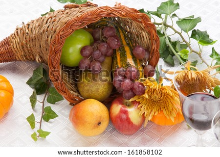 Horn of plenty full of fresh fruits - apples, pears, and grapes - on fall festival decorated table with pumpkins, flowers, and red wine