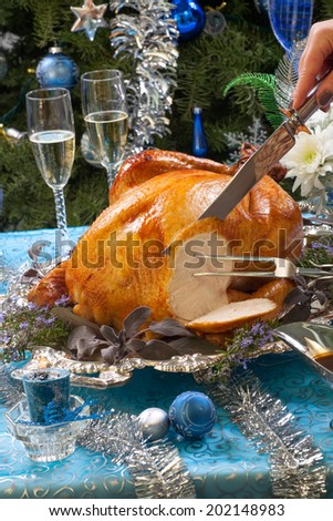 Carving roasted turkey garnished with herbs on blue Christmas decorations, and champagne. Christmas tree as background.