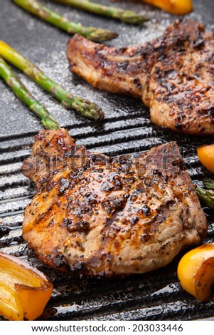 Juicy pork chops are grilled on griddle with asparagus and bell pepper. Backyard grilling for summer picnic.