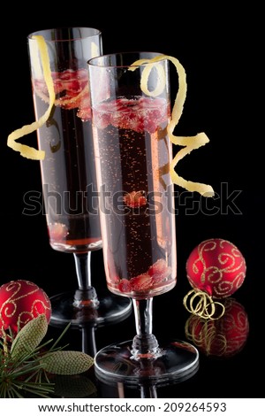 Closeup of glasses of pomegranate champagne cocktail, garnished with lemon twist, and Christmas ornaments on black background.