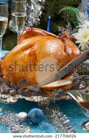 Carving roasted turkey garnished with herbs on blue Christmas decorations, and champagne. Christmas tree as background.