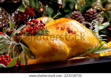 Roasted turkey garnished with sage, rosemary, and red berries in a tray prepared for Christmas dinner. Holiday table, candles and Christmas tree with ornaments.