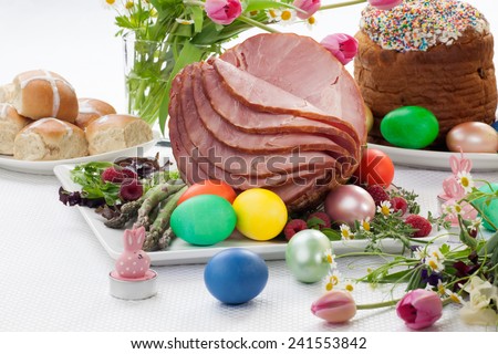 Whole baked honey sliced ham with fresh raspberry, asparagus, dyed Ester eggs, Easter cake, and cross buns. Spring flowers.