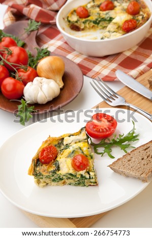 Closeup of plate with one piece of fresh made frittata, bread, and tomatoes. Pan with frittata with baby kale, sundried tomatoes, and goat cheese in background.
